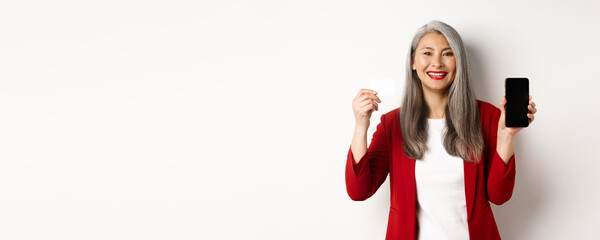 Senior asian businesswoman showing plastic credit card and blank smartphone screen, smiling at camera, white background