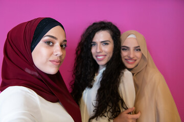 Wall Mural - Group of beautiful muslim women two of them in fashionable dress with hijab using mobile phone while taking selfie picture isolated on pink background representing modern islam fashion technology 