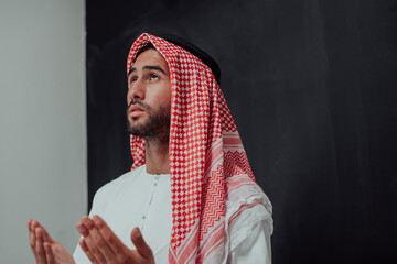 Wall Mural - Arabian man in traditional clothes making traditional prayer to God, keeps hands in praying gesture in front of black chalkboard representing modern islam fashion and ramadan kareem concept