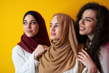 Wall Mural - Group portrait of beautiful Muslim women two of them in a fashionable dress with hijab isolated on a yellow background