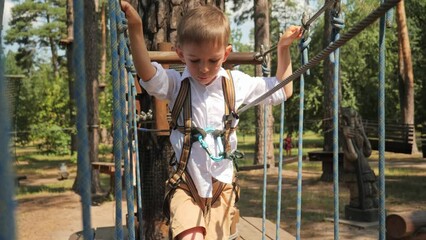 Wall Mural - Little boy holding rope while crossing bridge strung between two trees in park. Active childhood, healthy lifestyle, kids playing outdoors, children in nature