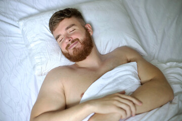 Wall Mural - Handsome young calm sleepy bearded man with beard is sleeping well on side in bed in bedroom on pillow with his eyes closed in the morning, resting. Good healthy dream, rest. White linens. Top view.