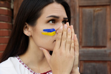 Sad young woman with drawing of Ukrainian flag on face near wooden door, closeup