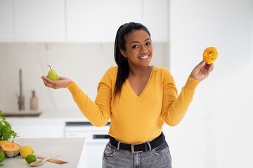 Wall Mural - Smiling pretty young african american female hold pear and donut at table with vegetables