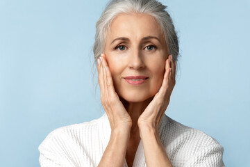 A beautiful elderly woman with perfect skin and gray hair touches her face with her hands. A mature woman in a white robe is nursing herself after a shower