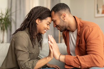 Wall Mural - Romantic african american spouses holding their hands, touching foreheads, bonding while sitting on sofa