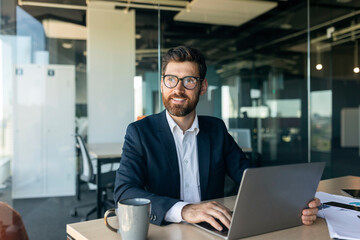 Mature male entrepreneur using laptop and thinking, looking aside while sitting at workplace in office, free space