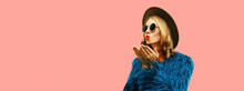 Fashionable Portrait Of Stylish Young Woman, Female Model Blowing Her Lips With Red Lipstick Sending Sweet Air Kiss Wearing Blue Faux Fur, Round Hat On Pink Background