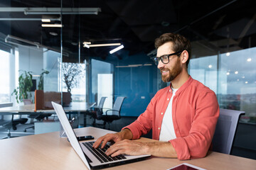 Successful smiling man working inside office with laptop, businessman in red shirt smiling and typing on keyboard in glasses, programmer working software for program.