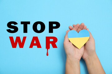 Poster - Stop War in Ukraine. Woman holding paper heart in colors of Ukrainian flag on light blue background, closeup