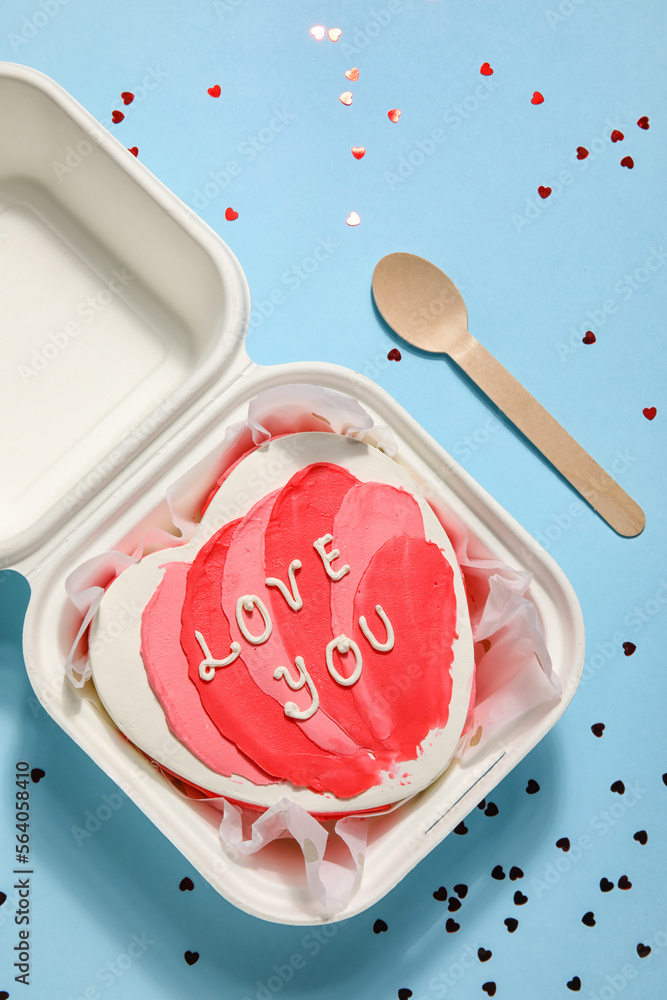 Plastic lunch box with heart-shaped bento cake and spoon on blue background. Valentine's Day celebration