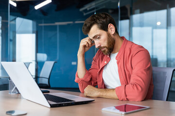 Frustrated businessman depressed at workplace working on laptop, man in shirt upset and sad displeased with bad work results and achievement inside office.