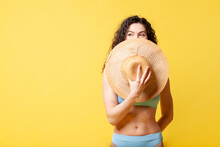 Young Slender Beautiful Brunette Girl In A Blue Swimsuit And With Curly Hair Holds A Straw Hat On A Yellow Background