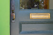 A dark green wooden door with half glass pane window, and a wooden panel. The vintage door has a brass door handle, a keyhole, and a letter plate. The trim of the building is vibrant green color. 