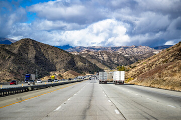 Big rigs semi trucks with semi trailers transporting different industrial cargo driving on the wide divided mountain highway road in California