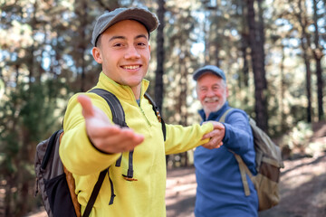 Wall Mural - Handsome young boy walks happily in the forest holding his grandfather's hand. Smiling multigenerational family enjoying freedom mountain and nature together. The new generation helps the old one