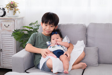 Asian elder brother and newborn baby sit in living room together, adorable infant sitting on boy lap with trust and comfortable. kid hold toddler in embrace with love and care. happy family concept.