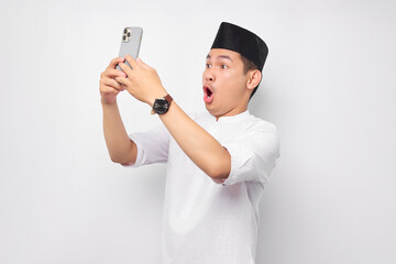 Surprised young Asian Muslim man looking at a smartphone screen and emotionally reacting to online news isolated on white background. People religious Islam lifestyle concept