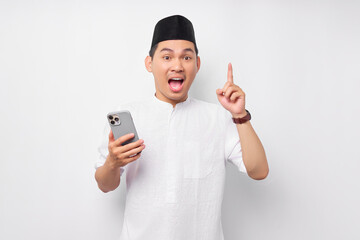 Excited young Asian Muslim man holding mobile phone, having creative idea, pointing his finger up on white background. People religious Islam lifestyle concept. celebration Ramadan and ied Mubarak