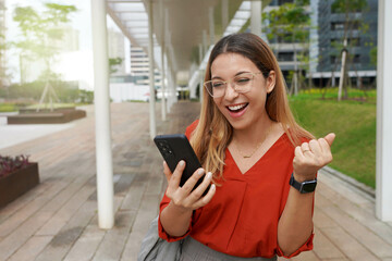 Canvas Print - Beautiful successful Brazilian female excited raised hand rejoicing with smartphone outside. Young woman watching at mobile phone celebrating good news from work in the morning.