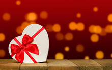 Valentine's Day Background With White Heart Shaped Present Box With Red Ribbon And Bow On Wooden Surface Table With Blurred Lights On Red Background