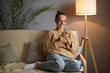 © sementsova321 - Photo of attractive smiling Caucasian woman wearing beige jumper and jeans sitting on cough, holding smart phone in hands, surfing internet, checking social networks.