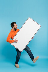 Wall Mural - full length of excited man holding huge template of smartphone on blue background.