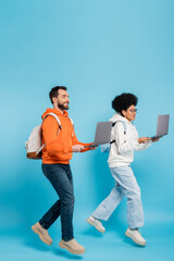 Canvas Print - full length of stylish multiethnic students with backpacks and laptops levitating on blue background.