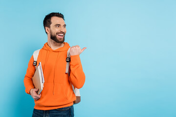 Wall Mural - cheerful bearded student with copybooks looking away and pointing with thumb isolated on blue.