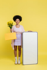 Wall Mural - full length of joyful african american woman with flowers and shopping bag near white template of mobile phone on yellow background.
