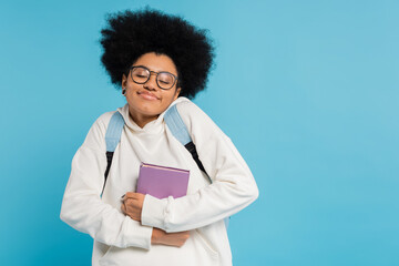 Wall Mural - pleased african american student in eyeglasses holding book while smiling with closed eyes isolated on blue.