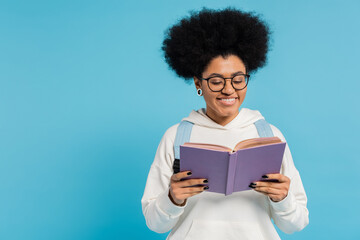 Wall Mural - curly and stylish african american student in eyeglasses smiling while reading book isolated on blue.