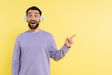 Wall Mural - amazed man in wireless headphones pointing with finger and looking at camera isolated on yellow.