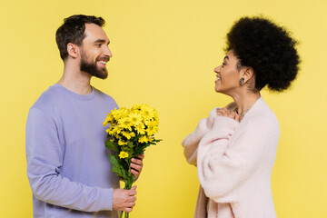 Wall Mural - happy bearded man holding bouquet near amazed african american woman isolated on yellow.