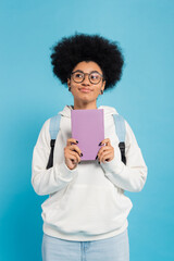 Wall Mural - dreamy and smiling african american student with backpack holding book and looking away isolated on blue.