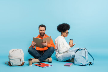 Wall Mural - bearded student looking in notebook near african american girlfriend sitting with smartphone on blue background.