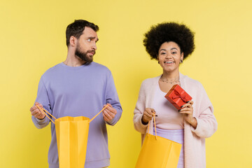 Wall Mural - happy african american woman holding gift box near offended man with shopping bag isolated on yellow.