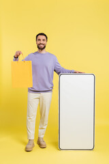 Wall Mural - full length of cheerful bearded man standing with shopping bag and huge template of smartphone on grey background.