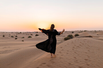 Back view of Beautiful mysterious woman in traditional arabic black long dress stands in the desert on sunset