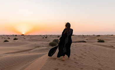 Back view of Beautiful mysterious woman in traditional arabic black long dress stands in the desert on sunset