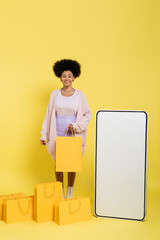 Wall Mural - cheerful african american woman smiling at camera near shopping bags and white template of mobile phone on yellow.
