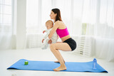 Fototapeta  - Young mom on maternity leave squats on yoga mat with baby in her arms. Sports at home after pregnancy