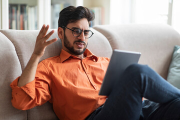 Wall Mural - Angry middle eastern man sitting on couch, using digital pad