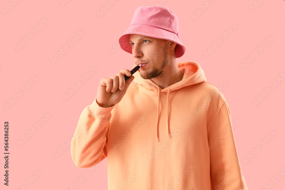 Young man in bucket hat smoking disposable electronic cigarette on pink background