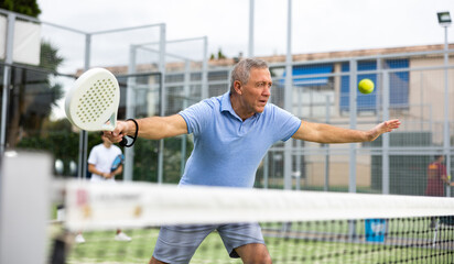 Wall Mural - Emotional mature male playing padel behind the net during match on tennis court in autumn