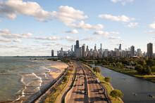 Downtown Chicago City Skyline Aerial Centered Over Traffic Along Lake Shore Drive Between South Lagoon And Lake Michigan On A Sunny Day With Fluffy White Clouds In A Blue Sky Above.