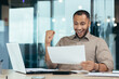 © Liubomir - Happy young african american businessman reading documents, letter, agreement, contract. Sitting at the table in the office, showing yes with his hand, celebrating.