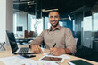 © Liubomir - A young African-American male accountant, auditor works in the office at a table with a laptop and documents. He holds a pen in his hands, looks at the camera, smiles