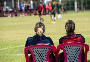 Moms sitting and watching their sons playing football in a school football tournament on a sideline with a sunny day. Sport, outdoor active, lifestyle, happy family and soccer mom concept.