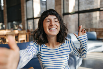 Wall Mural - Caucasian happy pretty woman staying with crossed fingers in bright room. Doing selfie on camera at cafe, wearing striped blue shirt. Different emotion concept